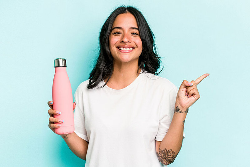 Junge hispanische Frau, die ein rosa Thermoskanns in der linken Hand hält, isoliert auf blauem Hintergrund, lächelnd und zur Seite zeigend.