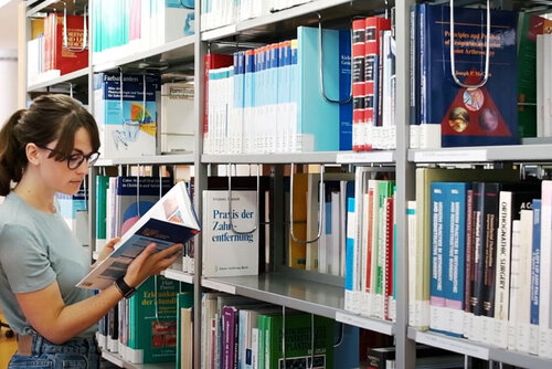 Eine Studierende in der Bibliothek. Sie steht vor einen Bücherregal und blättert in einem Buch.